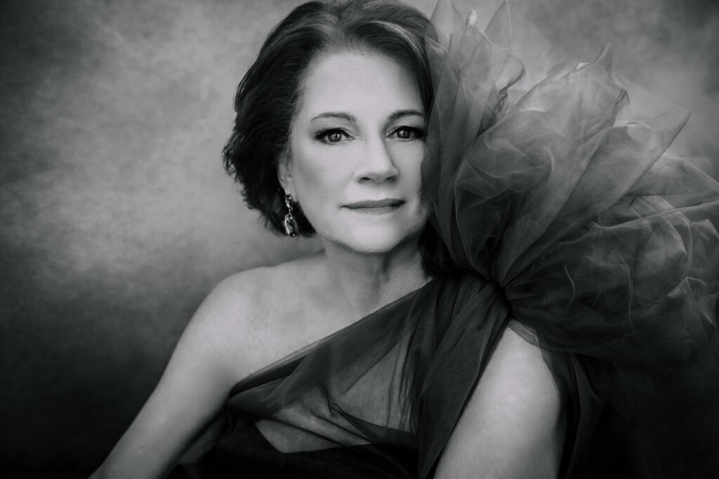 A woman with short dark hair wears a dramatic, tulle-ruffled dress and earrings, gazing confidently at the camera against a softly blurred background. The image is in black and white.