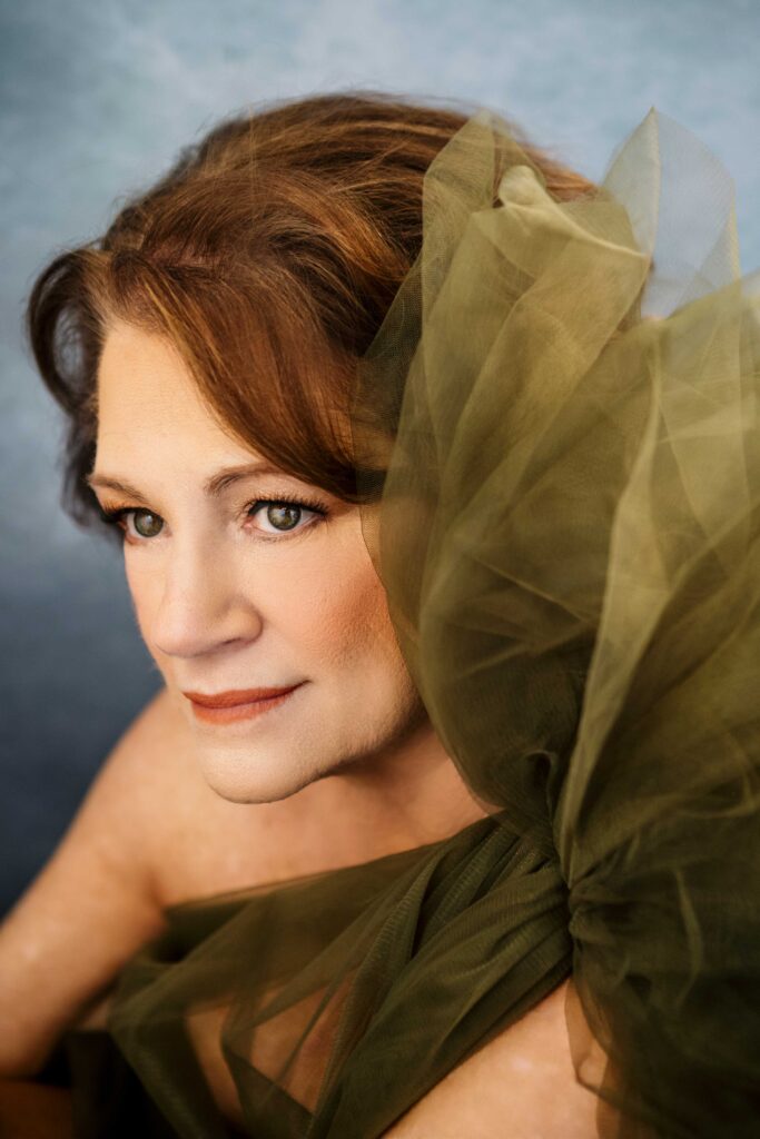 A woman with light skin and auburn hair gazes softly to the side. She wears a sheer, olive-green tulle garment with large, flowing fabric details on her shoulder, set against a muted blue background.