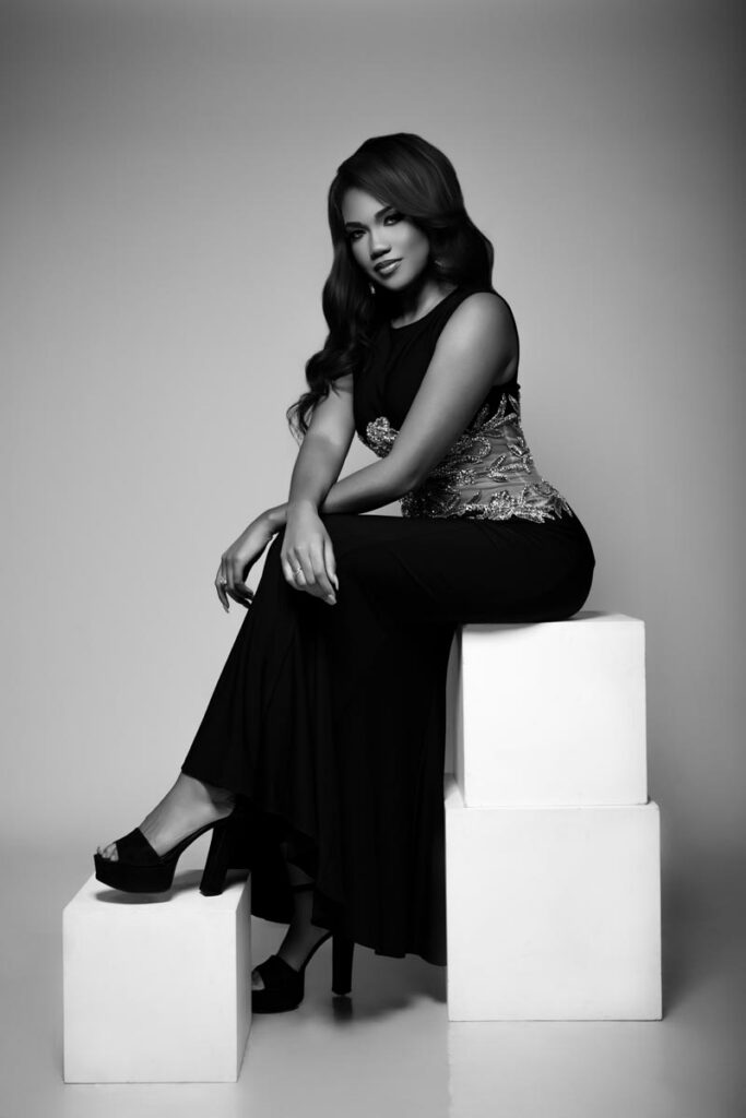 A woman in a long black dress and high heels sits elegantly on stacked white cubes, with one leg crossed over the other, against a plain, light background. The photo is in black and white.
