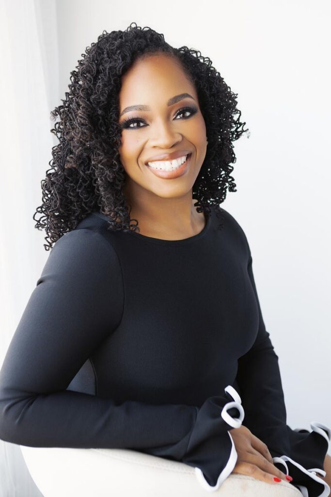 A woman with curly black hair, wearing a black long-sleeve top with white trim on the sleeves, sits and smiles at the camera against a bright, white background—perfect inspiration for what to wear for portraits Hagerstown MD.