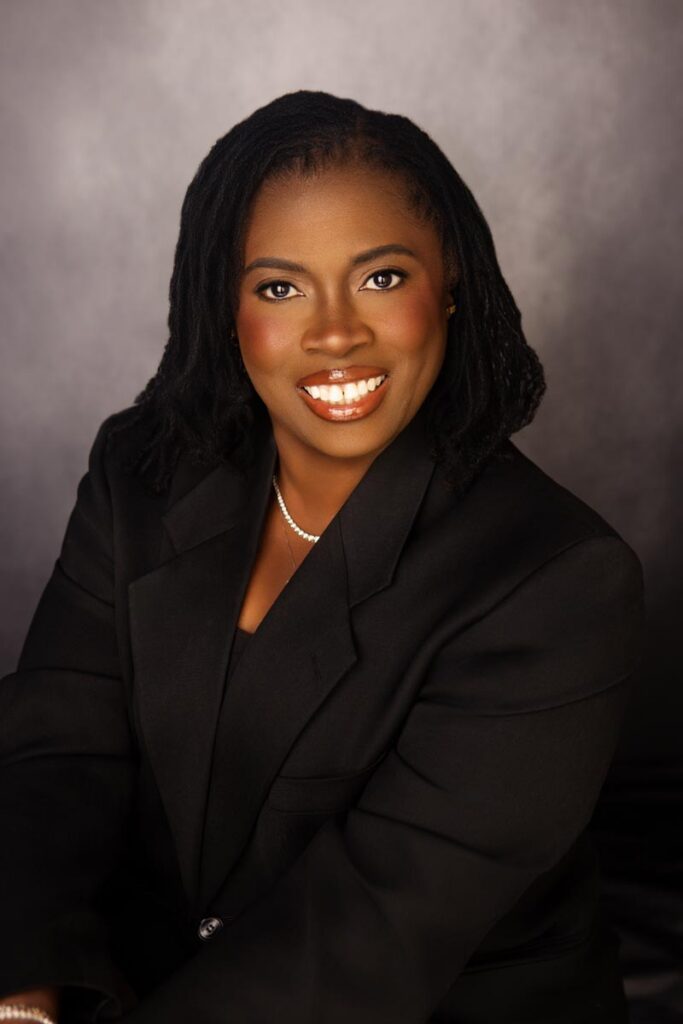 A woman sits wearing a black blazer and a pearl necklace smiling big in front of a gray textured background. This is one portrait outfit idea