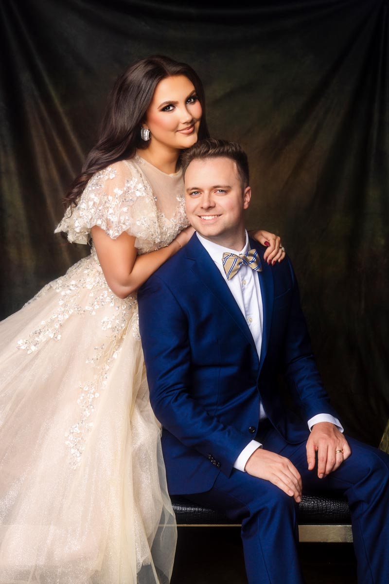 A woman in a light, beaded gown stands beside a seated man in a blue suit and bow tie. They smile warmly against a dark, textured backdrop.