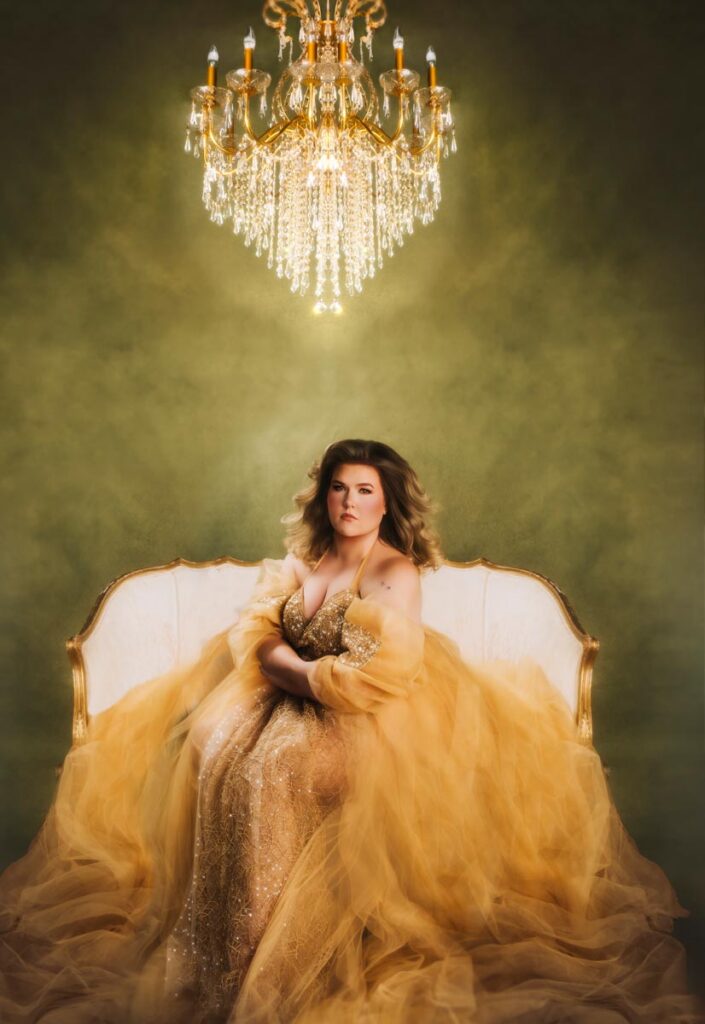 A woman wearing a large gold tulle gown with glitter accents sits on a white and gold sette underneath a large golden chandelier with a green background. Her styling for portraits was glamorous.