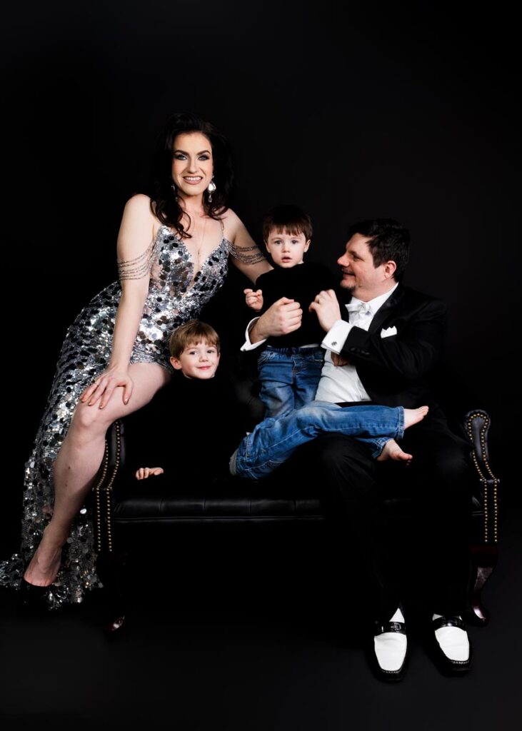 A woman in a sparkling silver dress, a man in a tuxedo, and two young boys in sweaters sit on a dark bench against a black background, smiling warmly captured perfectly by a talented Baltimore family photographer for a custom portrait photography experience.