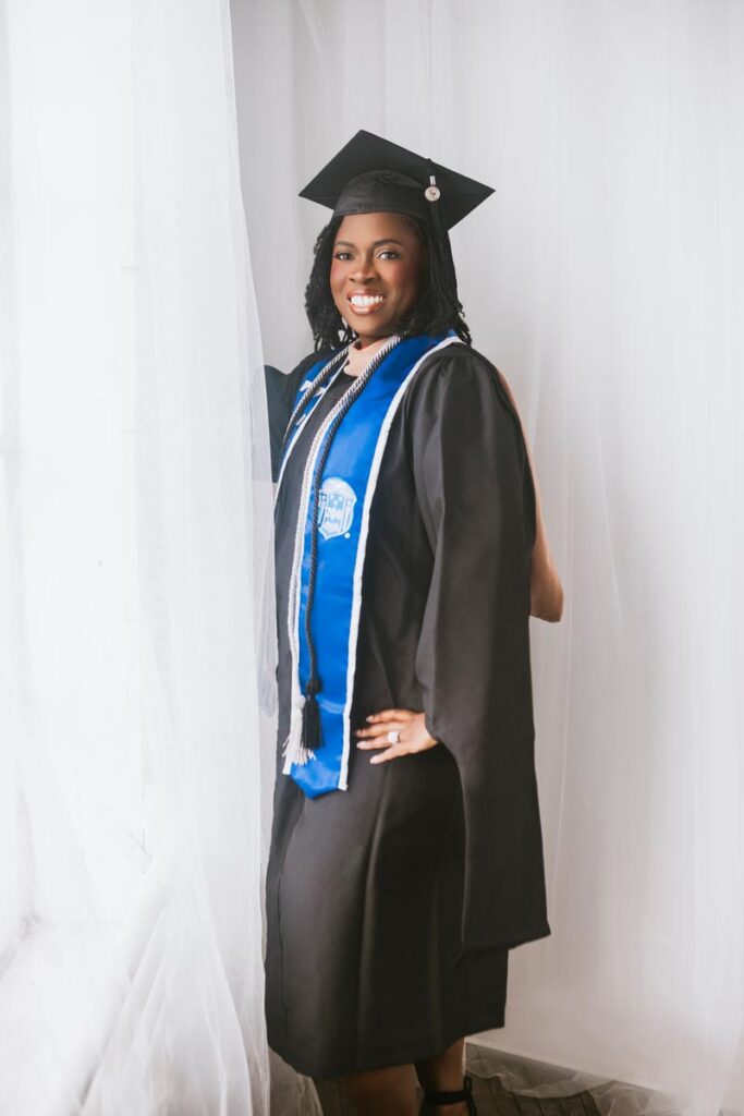 A woman wearing a graduation cap, gown, and stolls poses with her hand on her hip in front of a bright window and smiling.