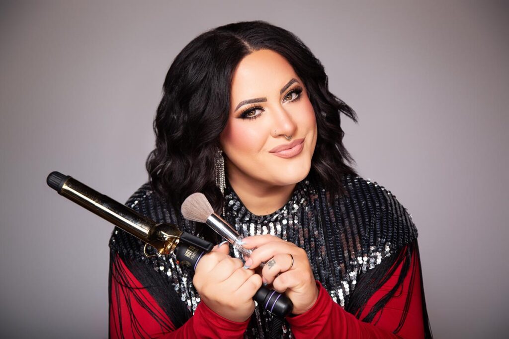A person with wavy, shoulder-length dark hair smiles while holding a curling iron and makeup brush—perfect for hair stylist branding photography. They wear a red and black sequined outfit with dramatic makeup against a neutral gray background.