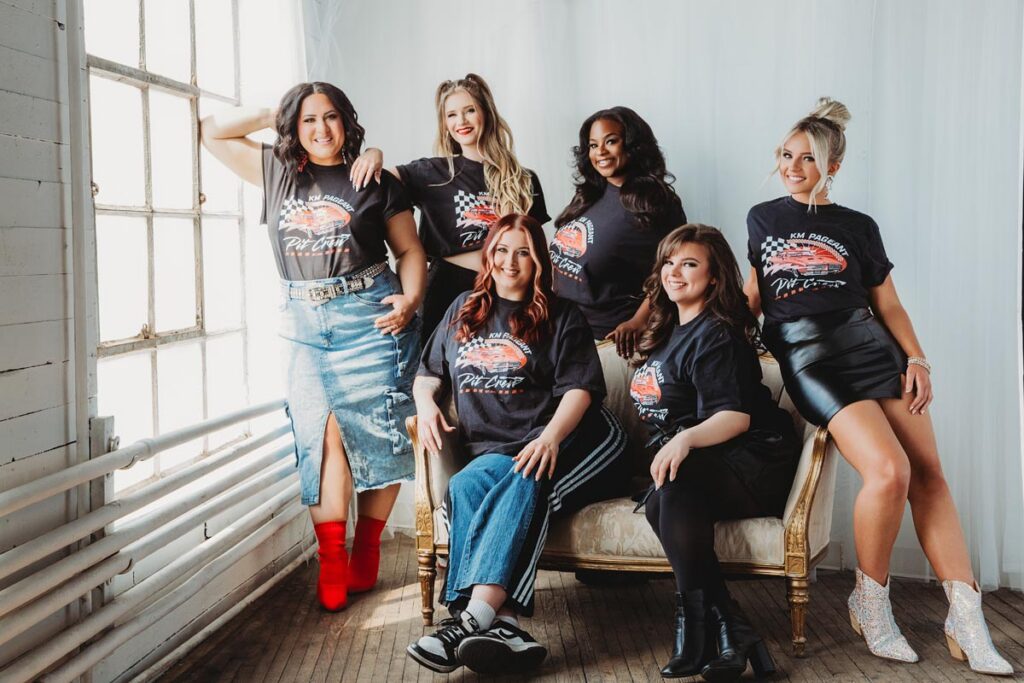 Six women pose together indoors for a branding photography session in Hagerstown, MD, all wearing matching black graphic t-shirts and various skirts or pants as they gather around a vintage sofa, smiling in natural light by a large window.