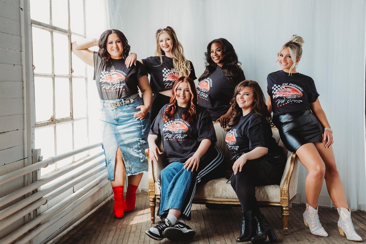 Six women pose together indoors for a branding photography session in Hagerstown, MD, all wearing matching black graphic t-shirts and various skirts or pants as they gather around a vintage sofa, smiling in natural light by a large window.