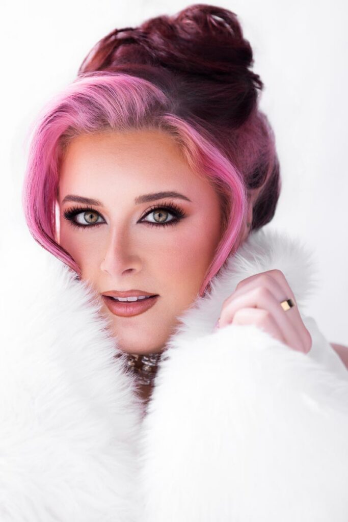 A person with pink and brown hair, glamorous makeup, and large jewelry poses in front of a white background under bright high key portrait lighting, wearing a white faux fur wrap while looking directly at the camera with a soft smile.