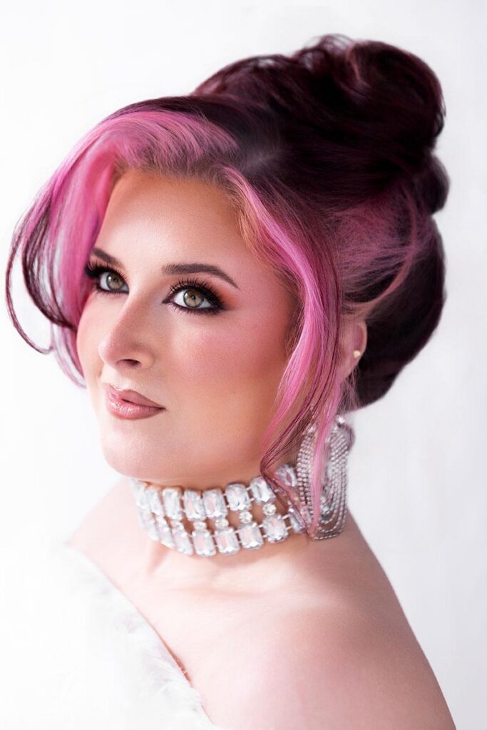 A person with pink and brown hair, glamorous makeup, and a large necklace poses in front of a white background under bright portrait lighting, wearing a white faux fur wrap while looking away from the camera.