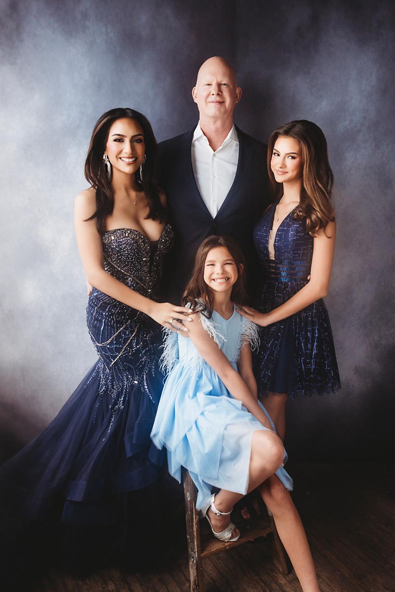 A family of four poses for a portrait against a dark background. Captured by a Portrait Photographer in Frederick, MD, an adult man in a suit stands behind three women and girls in elegant blue dresses as everyone looks toward the camera.