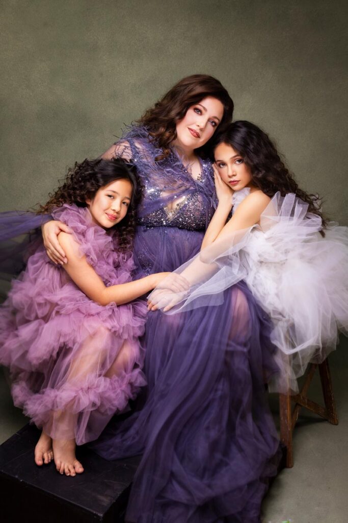 A woman in a purple gown sits between two young girls in ruffled tulle dresses, one in lavender and one in white. They are closely posed, sitting on stools against a muted backdrop, all with long, curly hair.