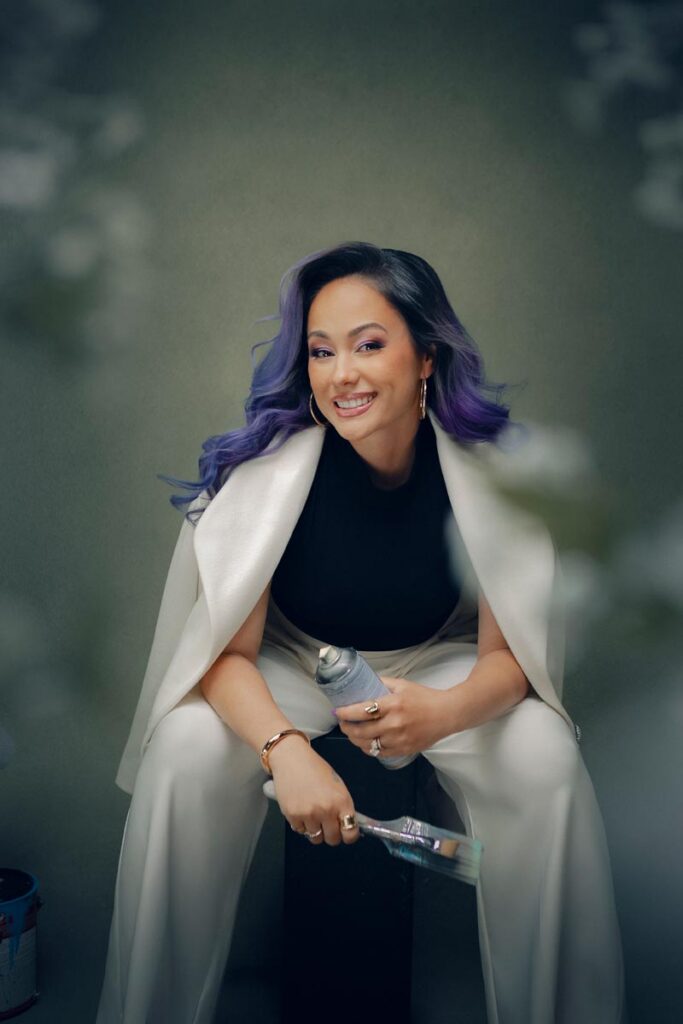 A woman with long wavy purple hair is seated, smiling, and holding a paintbrush and spray paint can. She wears a white suit over her shoulders and a black top—perfect for an artist branding photographer's creative portfolio, set against a blurred greenish background.