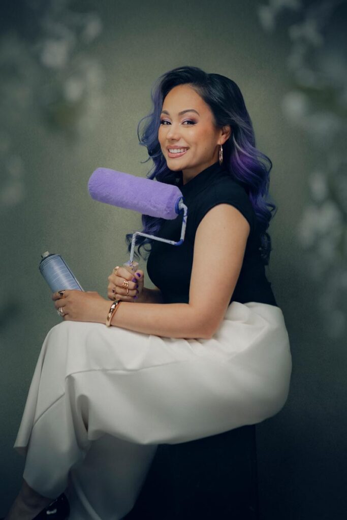 A smiling woman with long purple hair sits on a stool, holding a paint roller and a spray paint can. She wears a sleeveless black top and white pants, with a neutral background behind her.