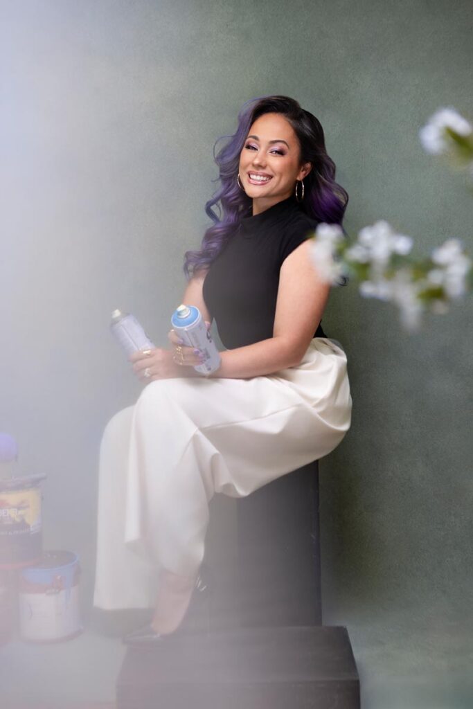 A smiling woman with long, wavy purple hair sits on a black box holding spray paint cans. She wears a black sleeveless top and white pants, with blurred flowers and paint cans visible in the foreground.