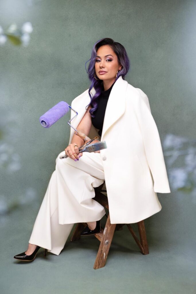 A woman with long purple hair sits confidently on a wooden stool, wearing a white suit draped over her shoulders and black heels, holding a paint roller with a purple roller head against a green backdrop.