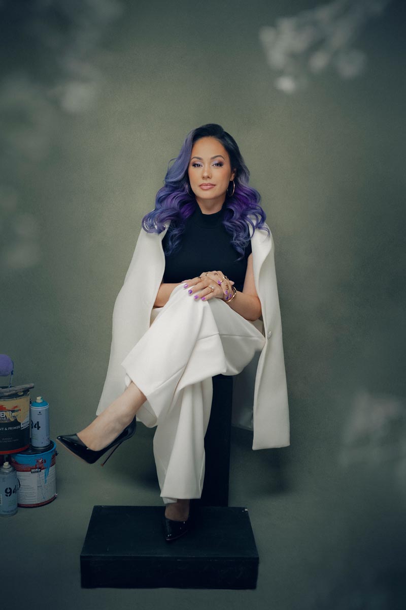 A woman with long wavy purple hair—reminiscent of painter Devona Stimpson's bold style—sits confidently on a black platform, white suit draped over her shoulders, with paint cans stacked beside her, embodying creative branding against a green backdrop.