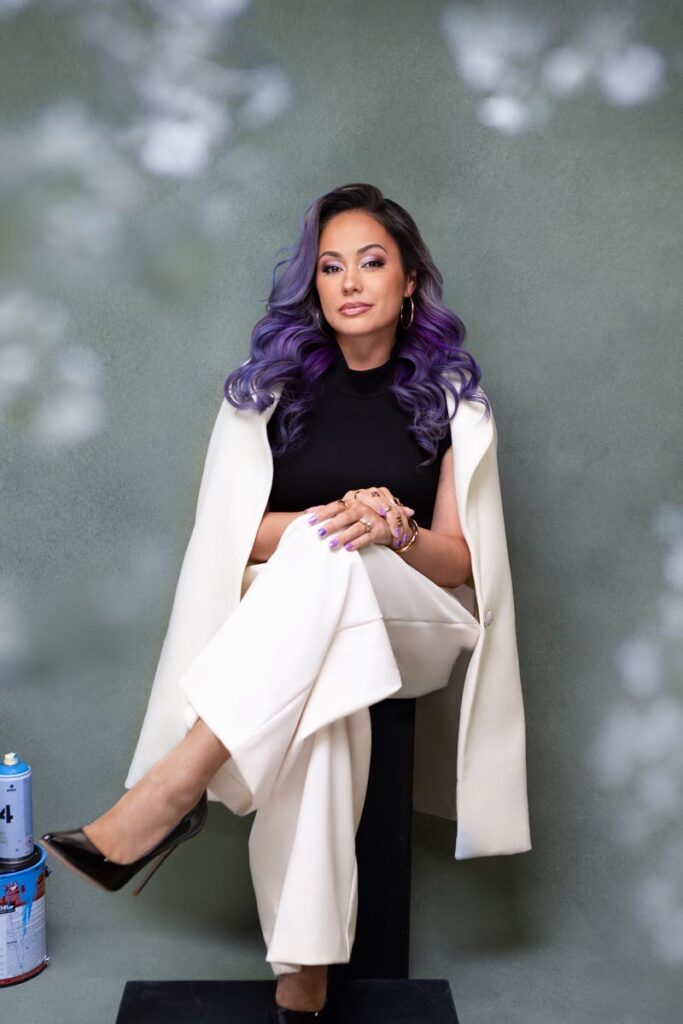 A woman with long wavy purple hair sits confidently on a black platform, white suit draped over her shoulders, with paint cans stacked beside her against a green backdrop.