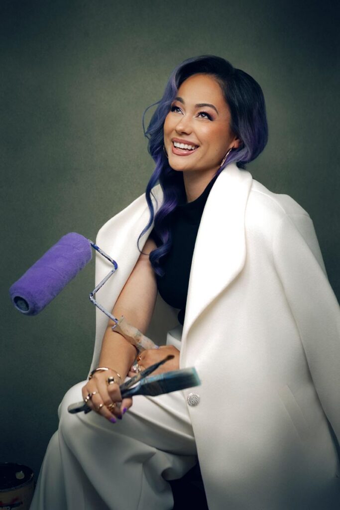A smiling woman with long, wavy purple hair wearing a white coat holds a purple paint roller and a paintbrush against a dark, plain background.