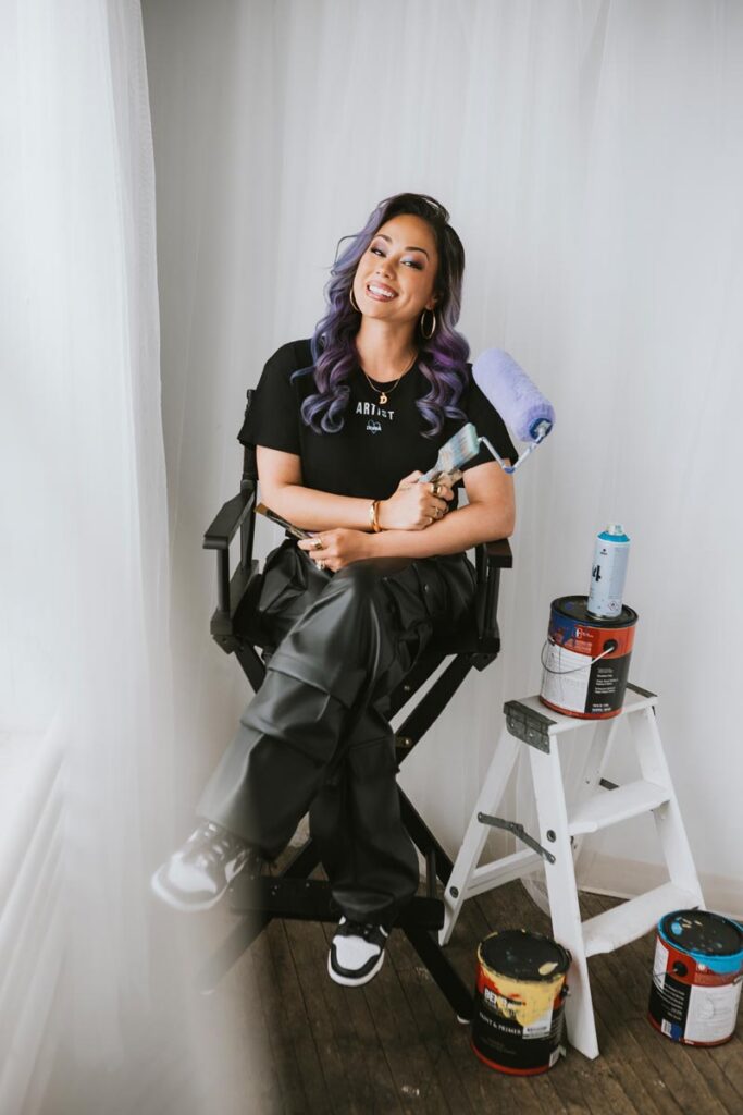 A woman with long wavy purple hair sits on a tall chair, smiling and holding a paint roller. She wears a black t-shirt and pants. Paint cans surround her, and a white step ladder is beside her. White curtains hang in the background.