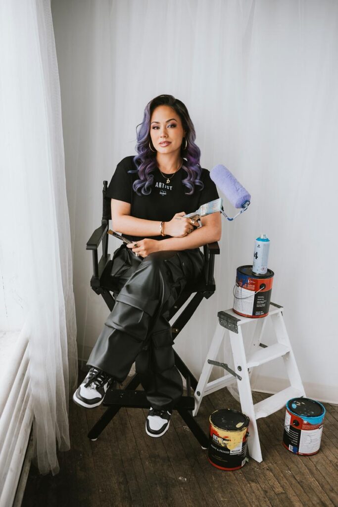A woman with purple hair sits on a tall black chair holding a paintbrush and roller. An artist branding photographer captures her in a bright room with sheer white curtains, paint cans, and a step ladder nearby.