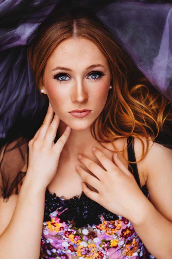 A young woman with long, wavy hair lies on a purple fabric. She gazes directly at the camera, with one hand touching her face and the other resting on her chest. She wears a colorful, floral-patterned top.