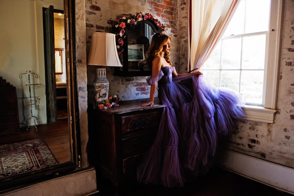 A woman in a flowing lavender gown sits on a vintage dresser by a window, gazing outside. The room has exposed brick walls, a decorative mirror with flowers, and elegant, antique furnishings.