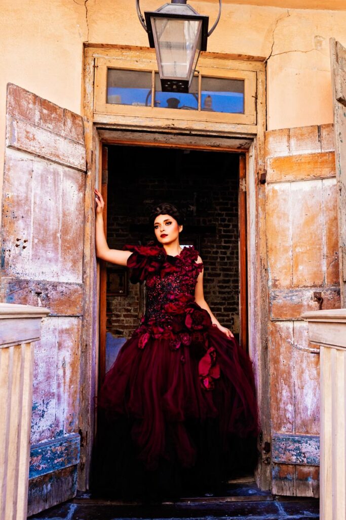 A woman in a dramatic, dark red gown with floral details stands against a rustic wooden door in a dimly lit room, gazing confidently at the camera. The background includes a brick wall and a partially visible window.
