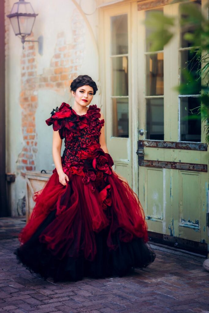 A woman in a dramatic, dark red gown with floral details stands against a rustic wooden door in a dimly lit room, gazing confidently at the camera. The background includes a brick wall