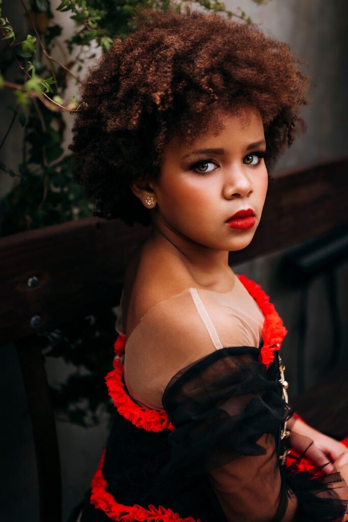 A young girl with curly hair and red lipstick sits on a wooden bench, wearing a black and red dress with sheer sleeves, looking thoughtfully at the camera. Green vines hang in the background.