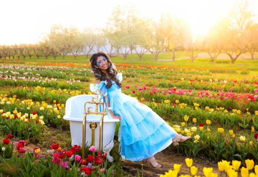 A woman in a blue, ruffled dress and flower crown sits on the edge of a white vintage bathtub in a colorful tulip field at sunset, smiling with one leg playfully raised amid blooming flowers.