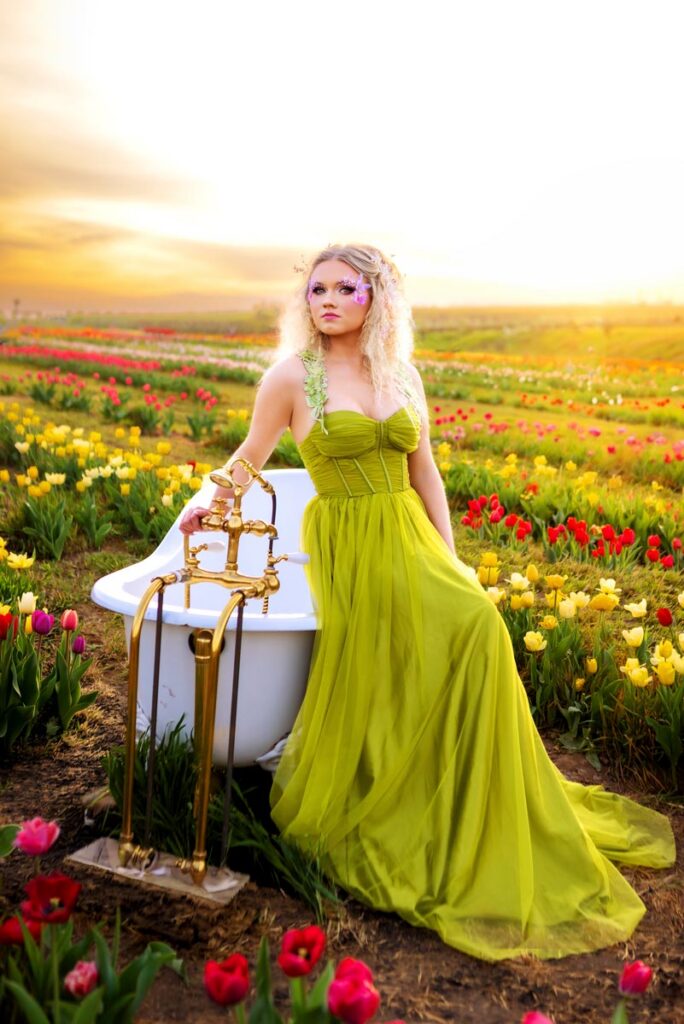 A woman in a long, flowing green dress sits on the edge of a white bathtub placed in a field of colorful tulips at sunset. She has wavy blonde hair, floral accessories, and looks off into the distance.