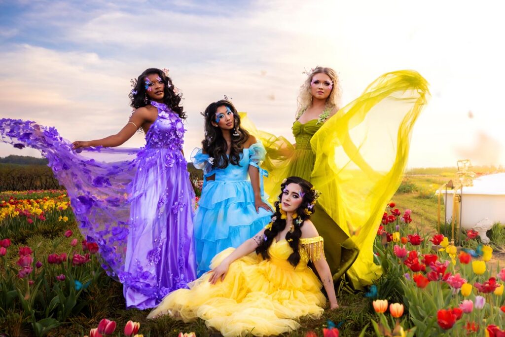 Four women in vibrant, flowing dresses—blue, yellow, green, and purple—pose in a field of colorful tulips at sunset. They have dramatic makeup and floral accessories, creating a whimsical, fairy-tale atmosphere.