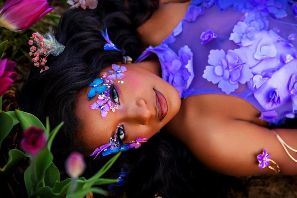 A woman with long dark hair lies amid flowers, wearing a purple dress with 3D floral designs. Her makeup features vivid purple and blue butterflies and petals on her face, creating a whimsical, enchanting look.