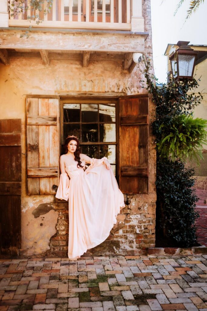 A woman in a long, flowing pale pink dress poses gracefully on a windowsill with wooden shutters, against a rustic brick and stucco building with vine and fern decorations.