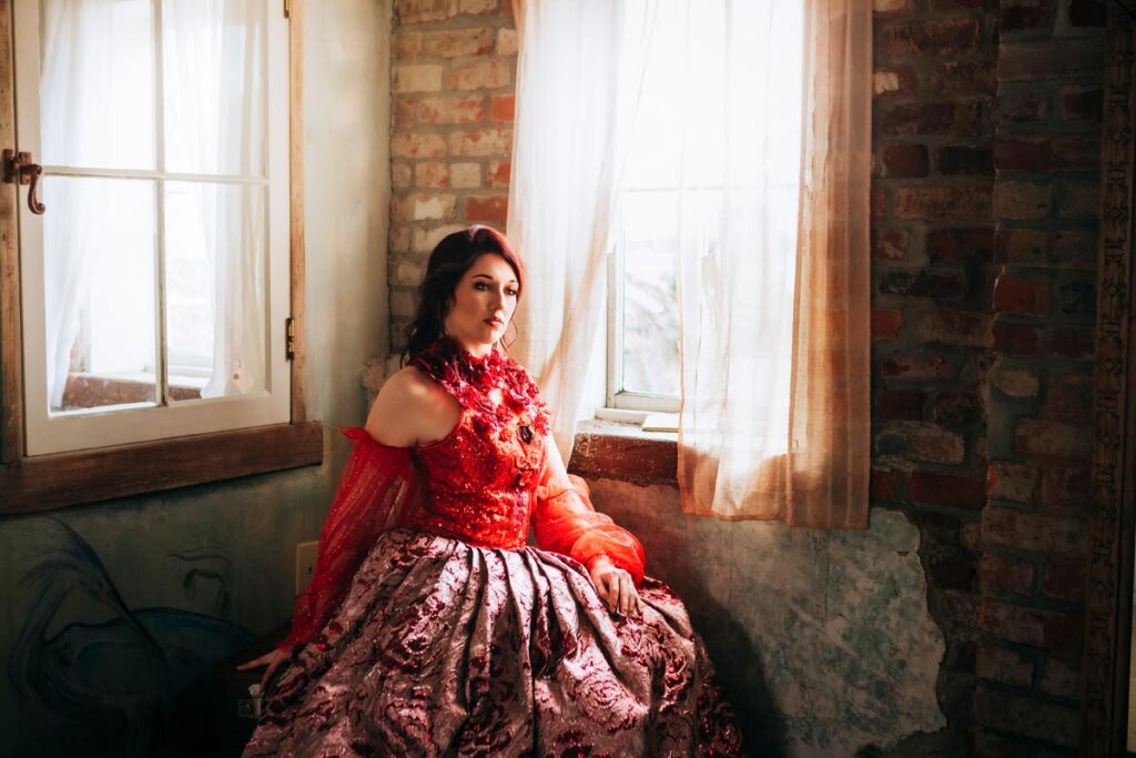 A woman in a red and pink ornate gown sits by a sunlit window with sheer curtains in a rustic room with exposed brick walls and peeling paint. She looks calmly at the camera.