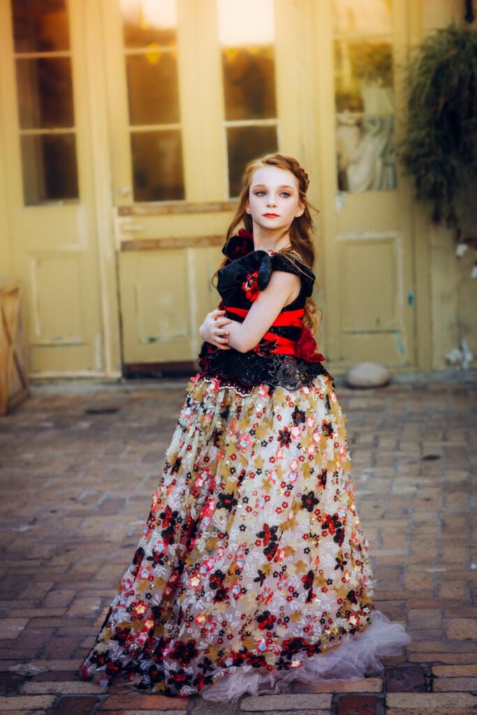 A young girl in a long, floral-patterned dress with a red and black bodice stands on a cobblestone path, looking over her shoulder in front of pale yellow double doors, with soft sunlight in the background.