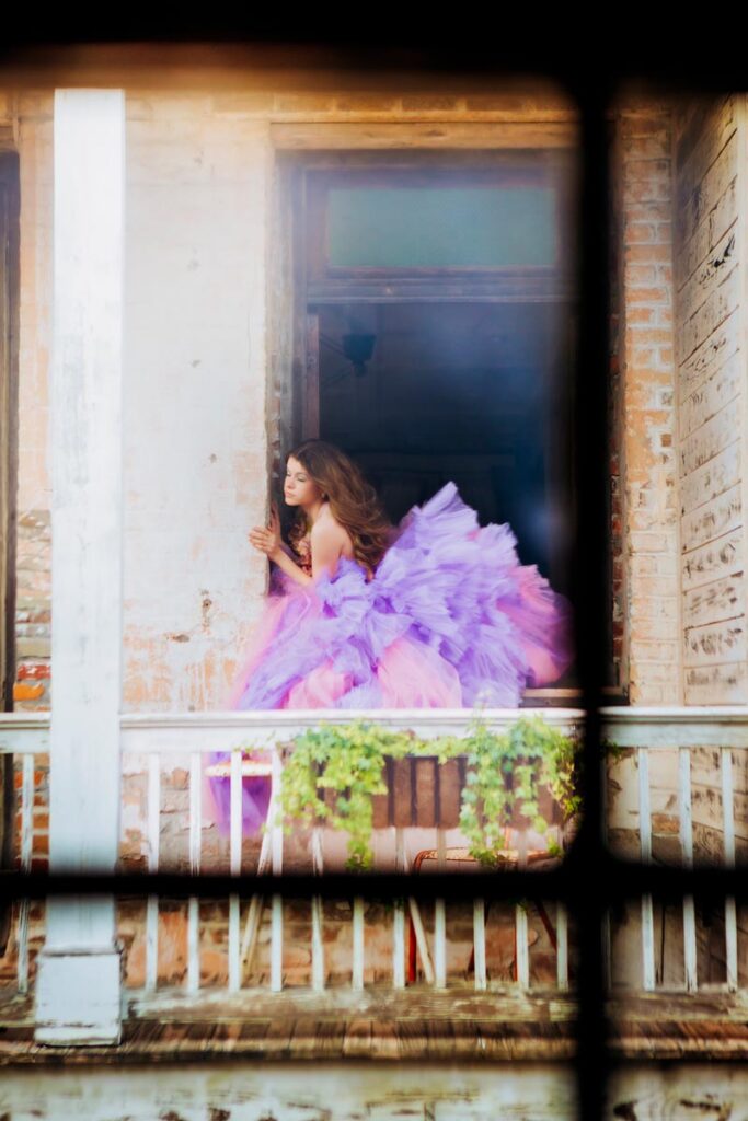 A woman in a voluminous purple and pink gown leans out of an old, weathered building’s window, with trailing greenery on the railing below. The photo is framed through a dark grid, adding a dreamy effect.