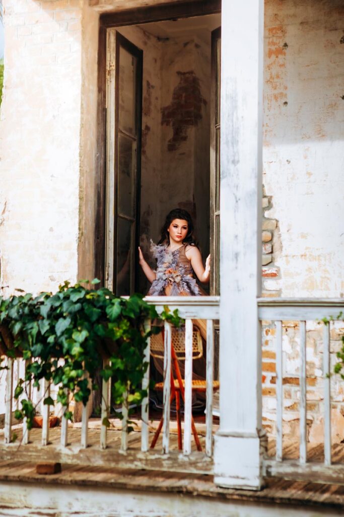 A woman in a feathery dress stands in an open doorway on a weathered, white-painted porch with green ivy growing on the railing. She looks outward, framed by peeling paint and rustic brick walls.