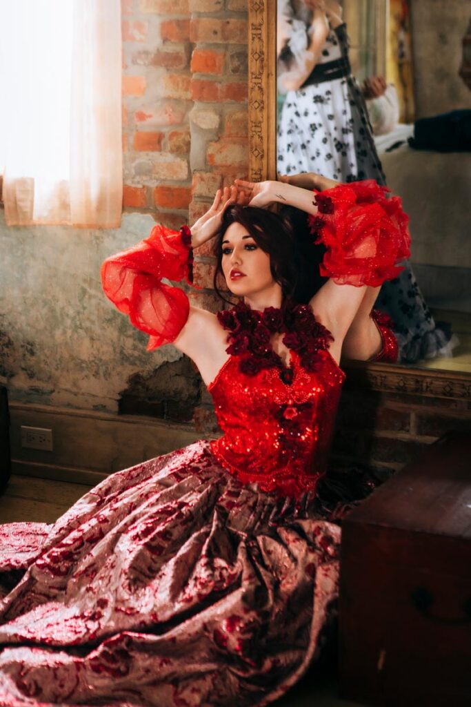 A woman in a red and pink ornate gown poses dramatically on the floor by a brick wall, with her arms raised and her reflection visible in a large mirror behind her. Warm light filters in from a nearby window.