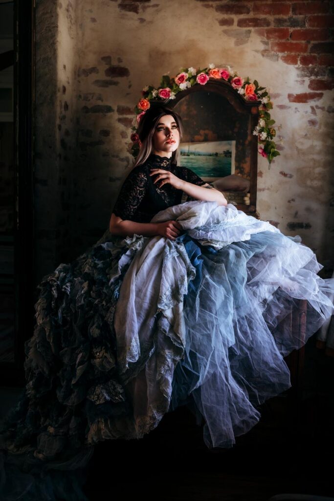 A woman in a dramatic, layered blue and white gown sits pensively by a rustic brick wall, with a floral wreath and a framed painting behind her, light casting a soft glow on her face and dress.