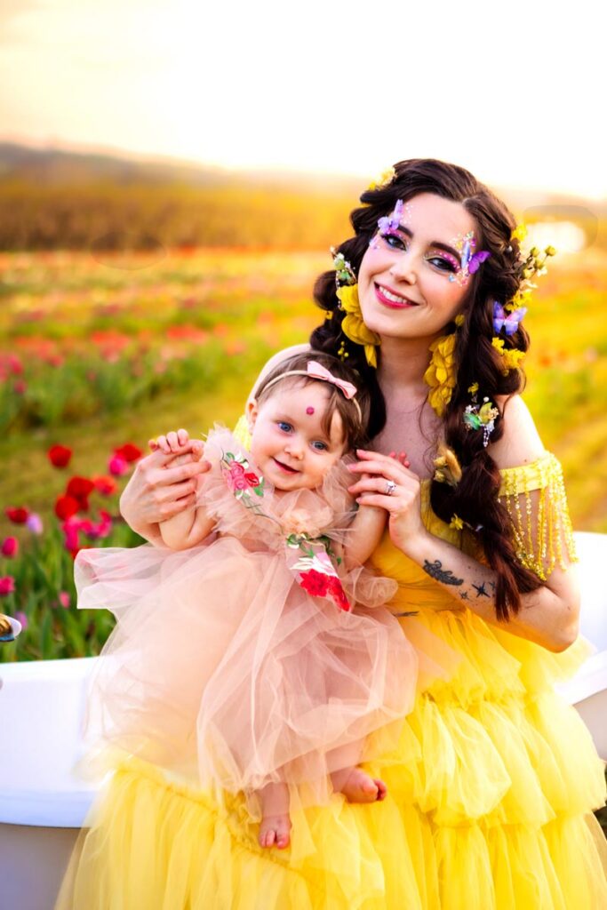 A smiling woman in a yellow dress with floral accessories and a baby in a pink dress sit together outdoors in a colorful flower field at sunset, both looking happy and festive.