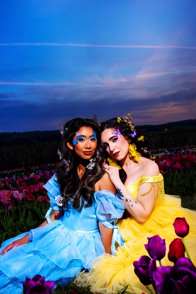 Two women in a vibrant flower field at dusk, one wearing a blue dress with blue floral makeup, the other in a yellow dress with yellow flowers in her hair. Both have dramatic makeup and sit closely together among tulips.