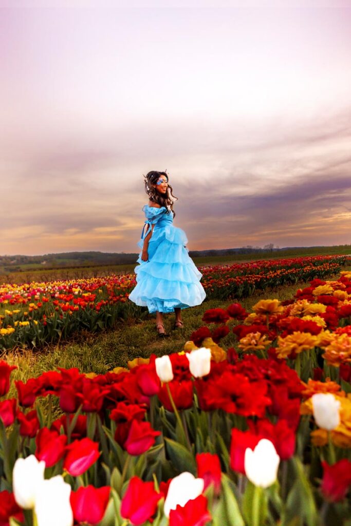A woman in a bright blue, ruffled dress stands in a vibrant field of red, yellow, and white tulips under a dramatic, cloudy sky at sunset or dusk.