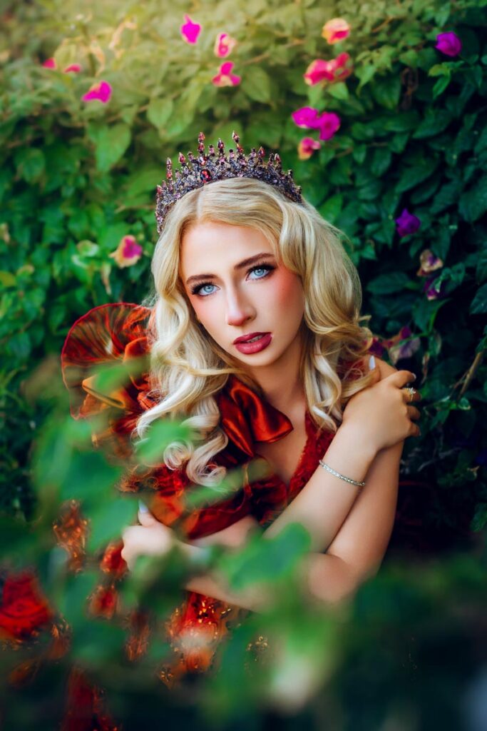 A blonde woman wearing a jeweled crown and a red dress poses among green foliage and pink flowers, gazing at the camera with her arms crossed over her chest.