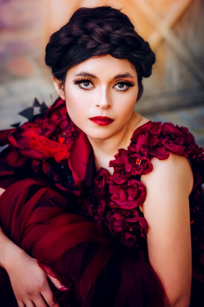 A woman in a dramatic, dark red gown with floral details stands against a rustic wooden door in a dimly lit room, gazing confidently at the camera. The background includes a brick wall and a partially visible window.