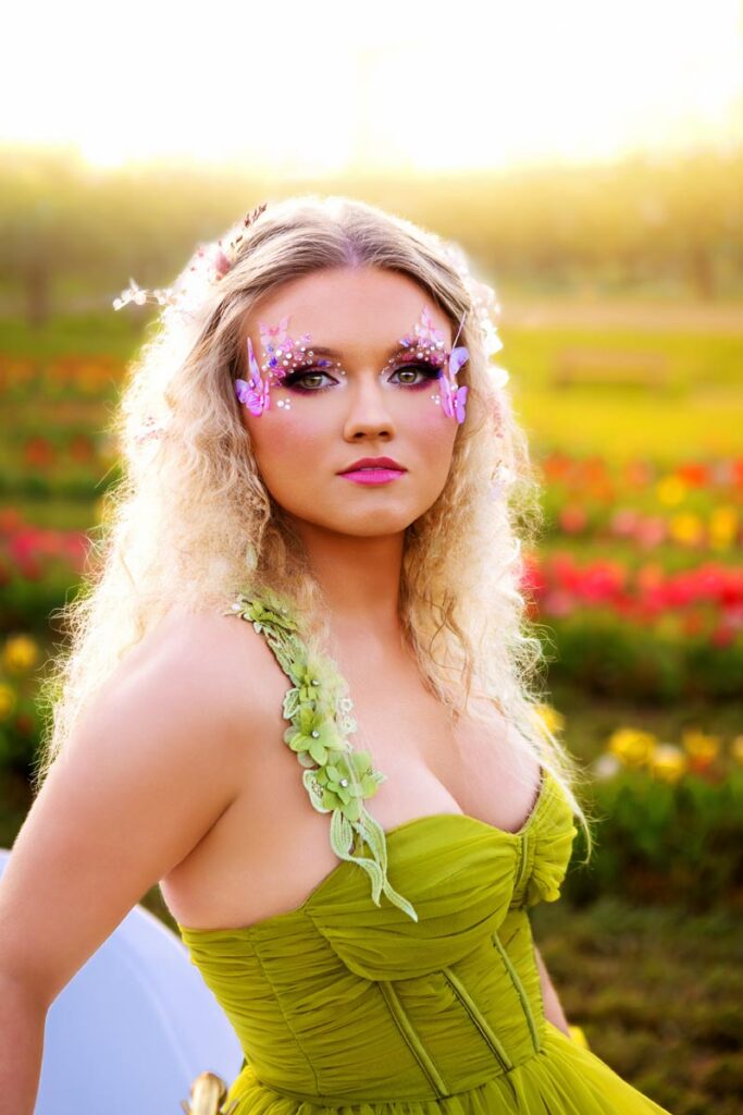 A woman in a long, flowing green dress sits on the edge of a white bathtub placed in a field of colorful tulips at sunset. She has wavy blonde hair, floral accessories, and looks off into the distance.