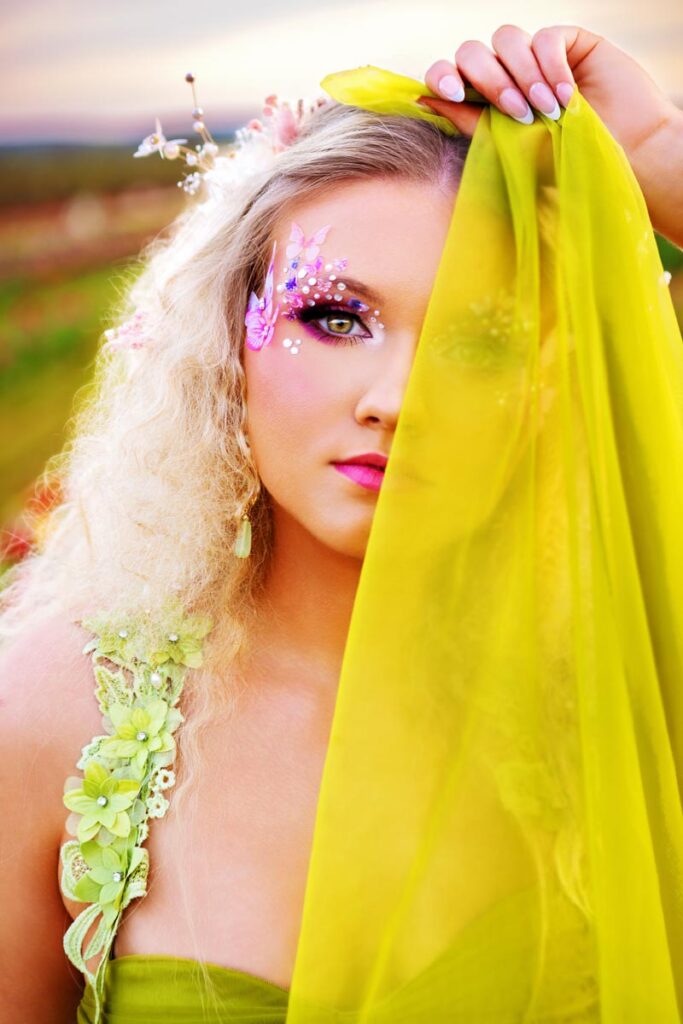A woman with curly blonde hair and floral makeup holds a sheer yellow fabric over half her face. She wears a green dress with flower details and stands outdoors with a blurred background.