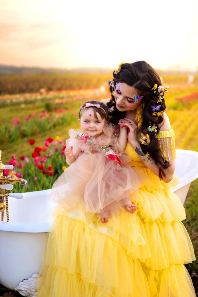 A woman and a baby, both wearing yellow and pink dresses, stand by a white bathtub in a flower field at sunset. The woman has flowers in her hair and looks lovingly at the smiling baby.