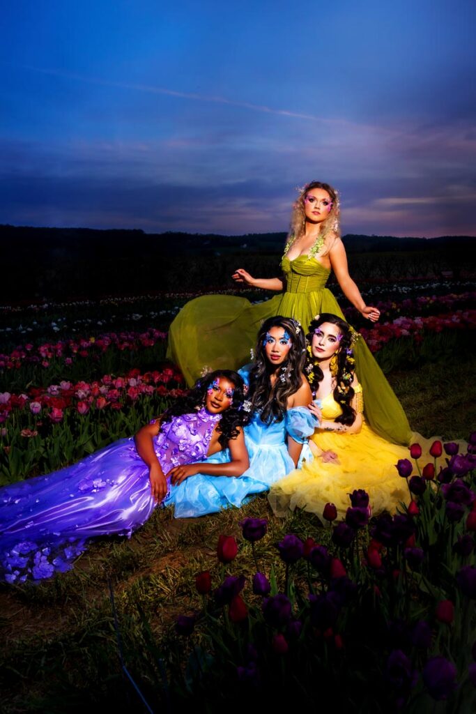 Four women in bright, flowing dresses (purple, blue, yellow, green) pose on a tulip field at dusk, with dramatic lighting and a vibrant sunset sky in the background.